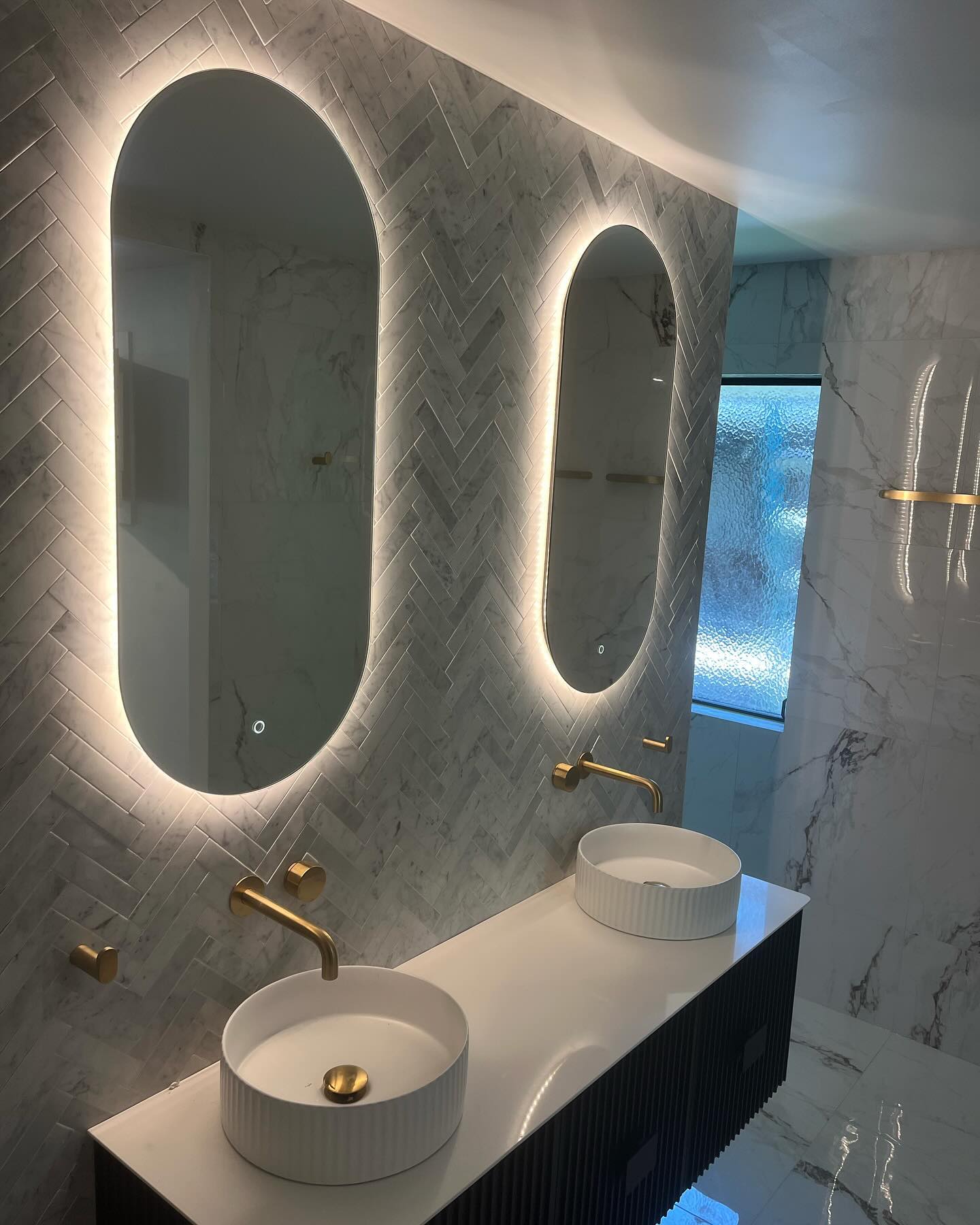 Bathroom with two oval LED backlit mirrors and a marble herringbone wall
